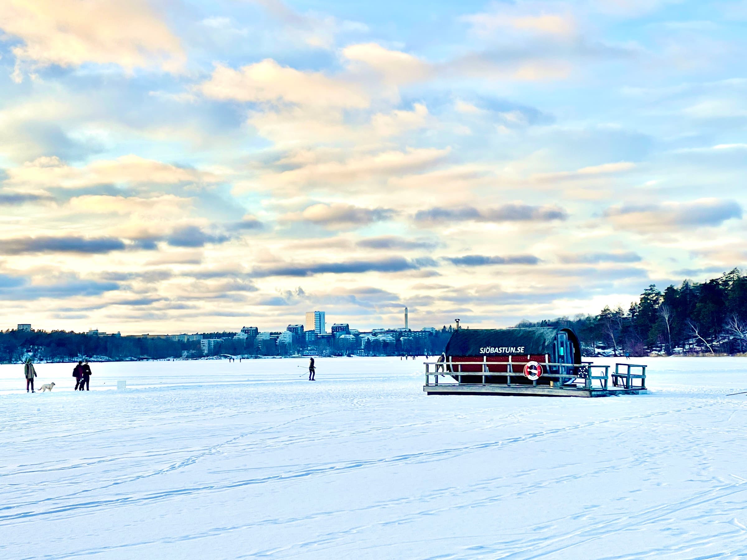 Bastuflotten på den snötäckta isen med skridskoåkare och promenerande en solig vinterdag